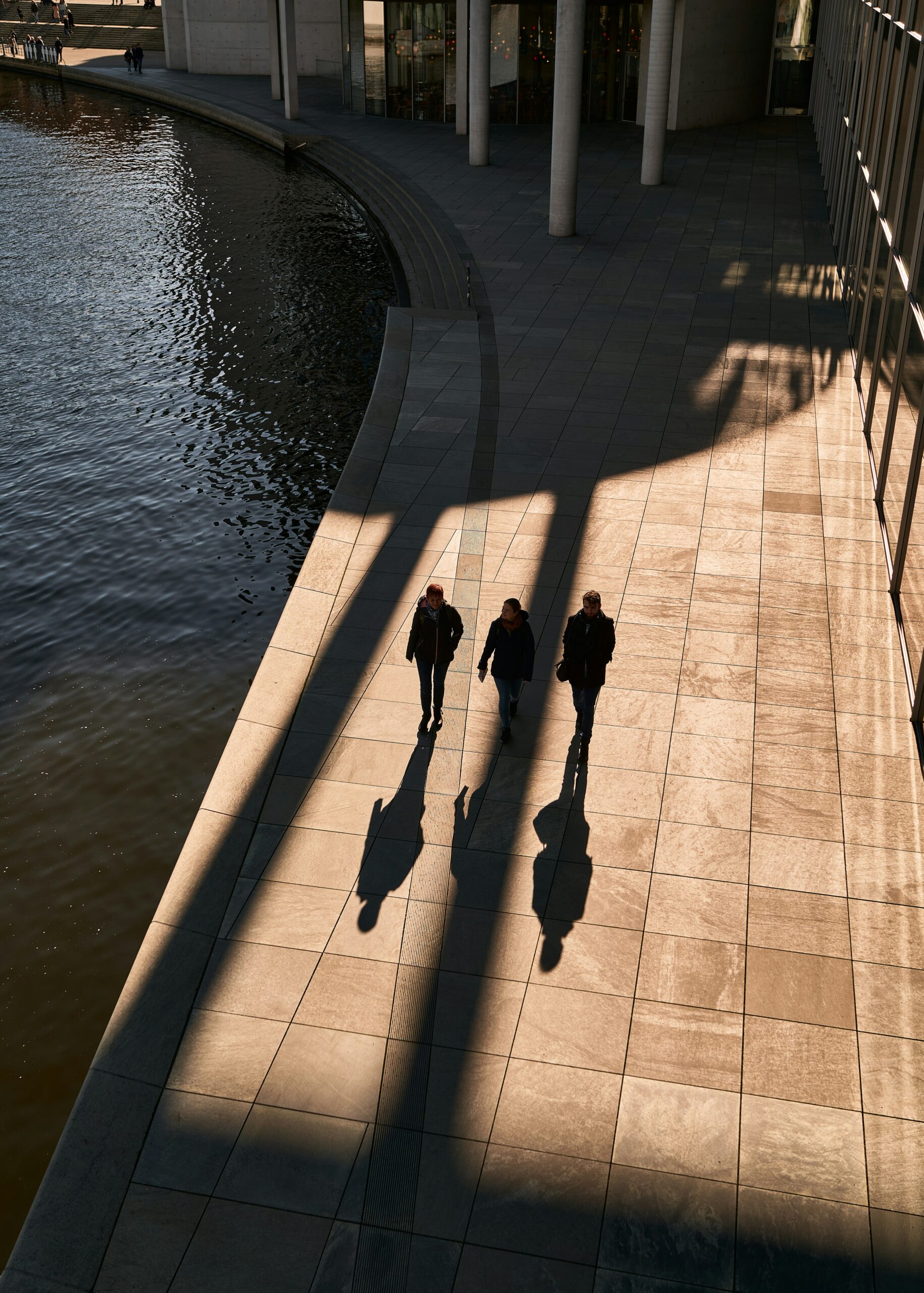 Three people walking beside a modern city building with long shadows cast across the pavement, symbolizing human connection and reflection in leadership -City Boss Coaching-Kellye Mazzoli